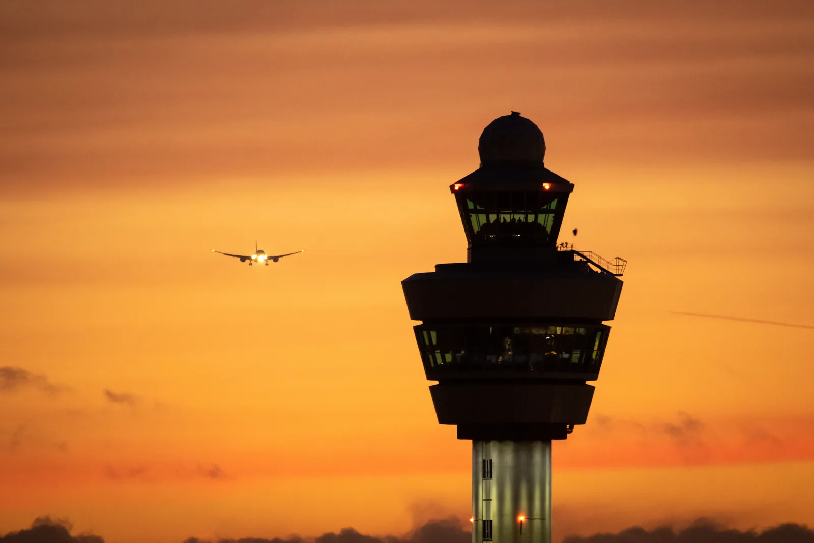 An airport control tower silhouetted against an orange sunset with a departing aircraft