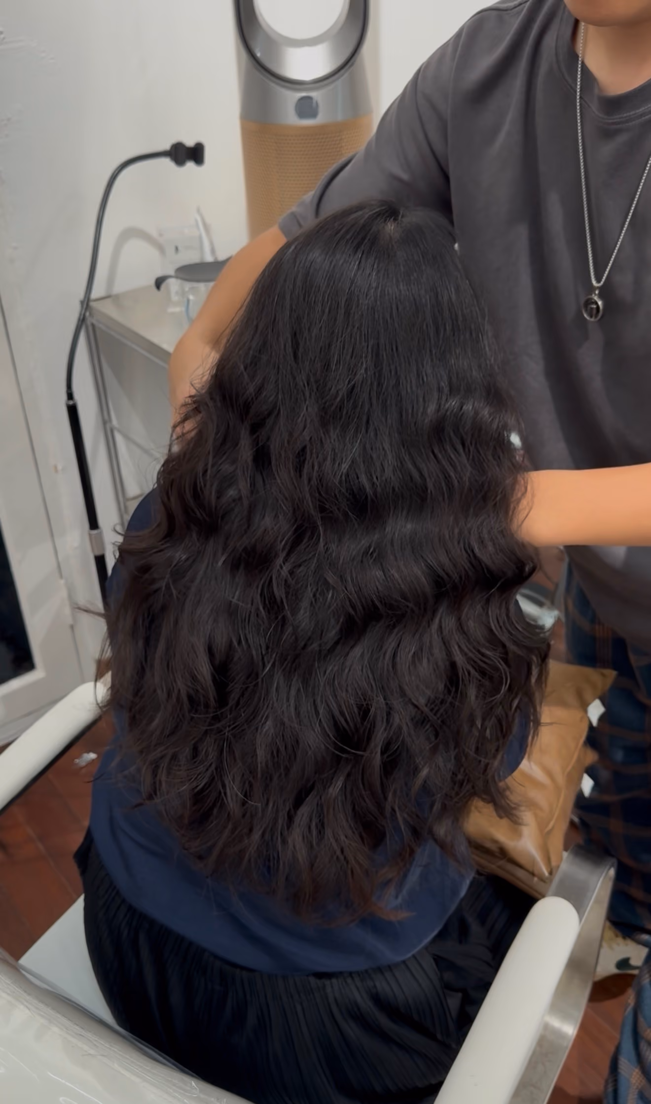 Person with long, wavy black hair seated in a salon chair with someone styling their hair.