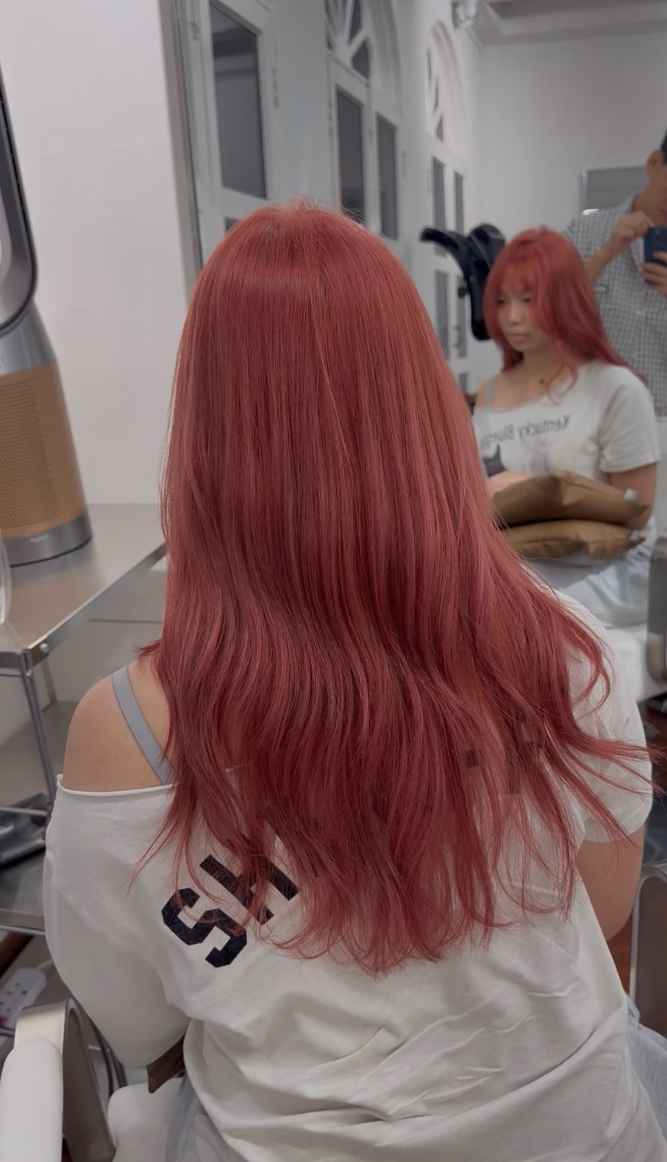 Person with long wavy rose red hair sitting in front of a mirror with their reflection visible.
