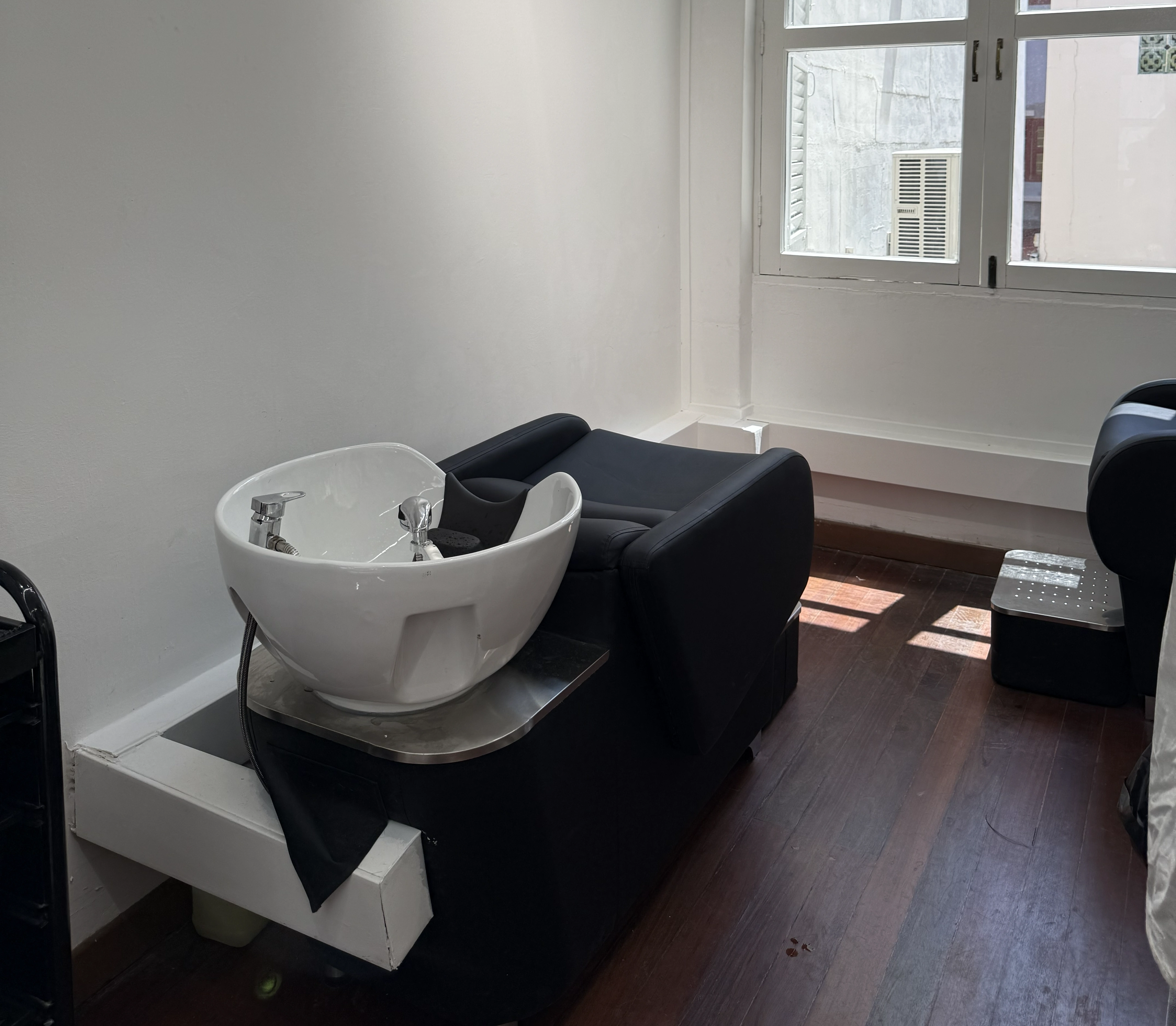 Black salon chair with white wash basin for hair washing in a room with wooden floor and window.