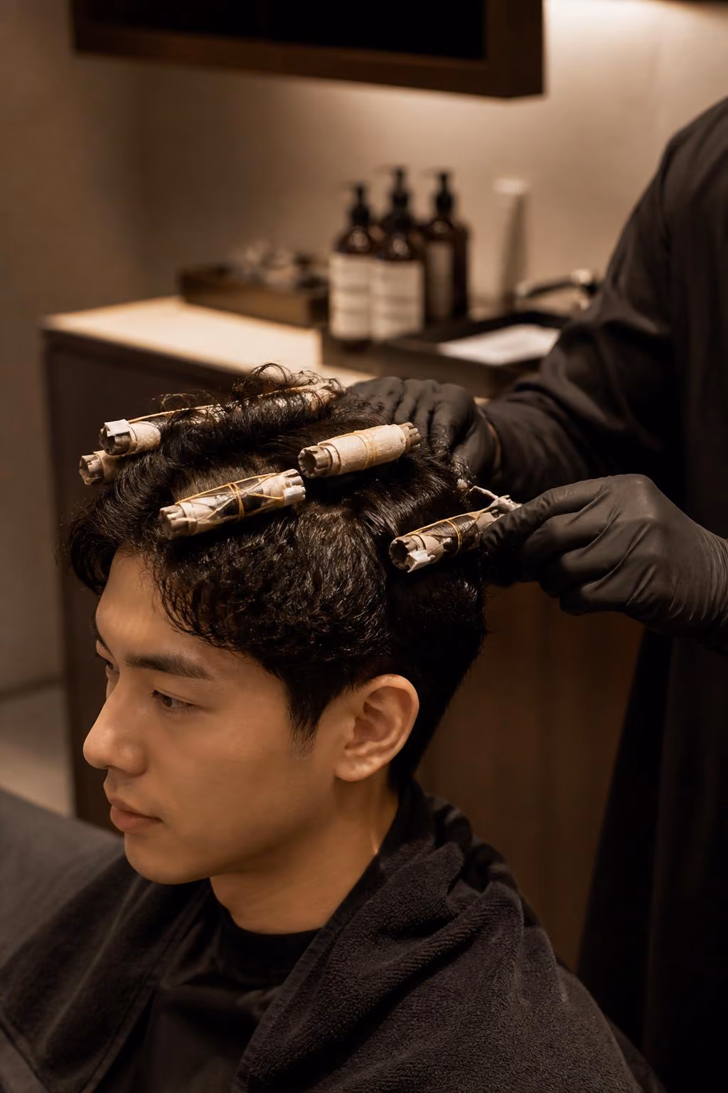 Man getting a perm with hair wrapped around rods by a stylist wearing black gloves in a salon.