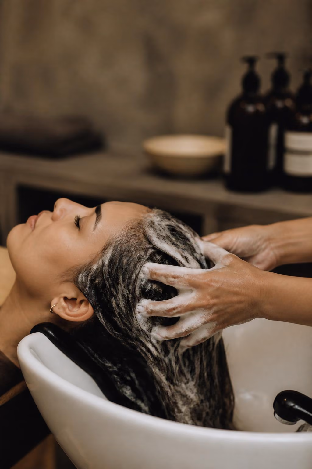 Woman with eyes closed reclining as hair is shampooed and massaged at a salon sink.
