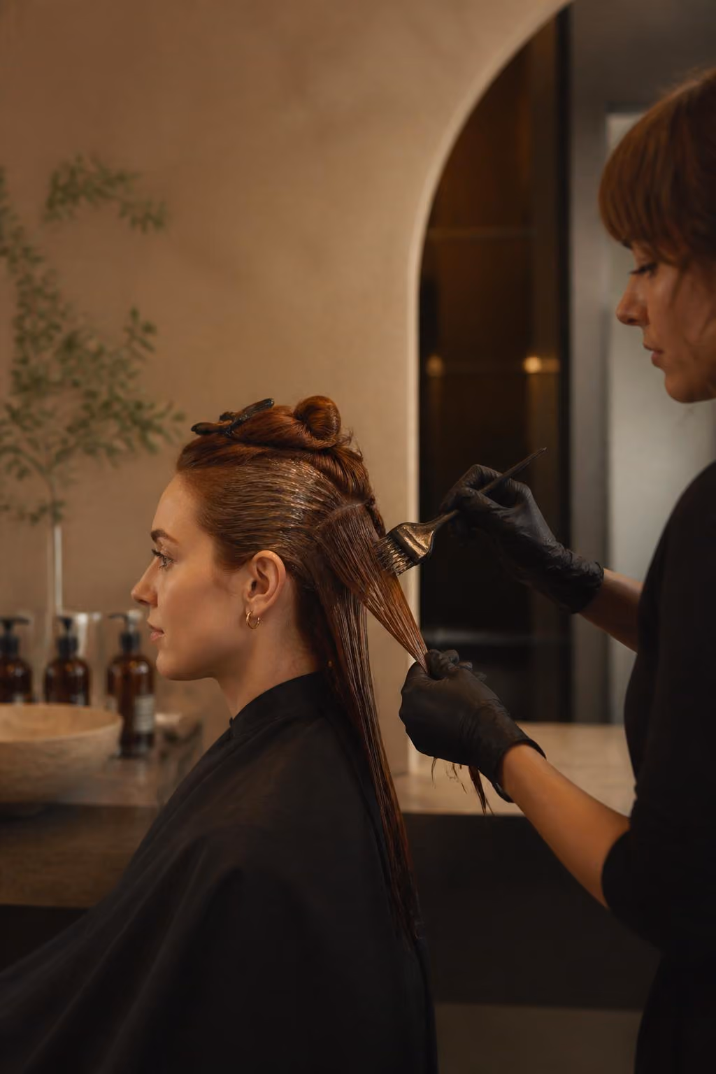 Hairdresser wearing black gloves applies hair dye to client's wet hair in a salon.