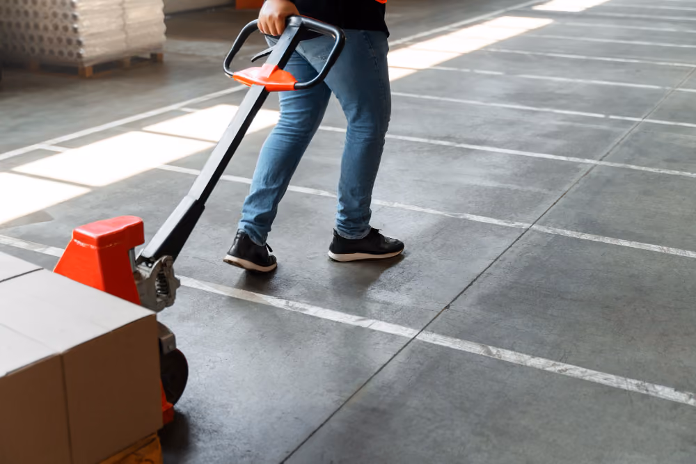 Person in jeans and sneakers pulling a red pallet jack with cardboard boxes on a warehouse floor.