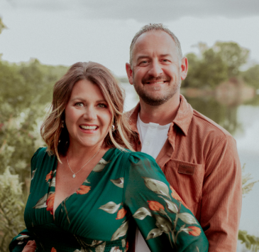 Smiling couple outdoors, woman in green floral dress and man in brown jacket by water.