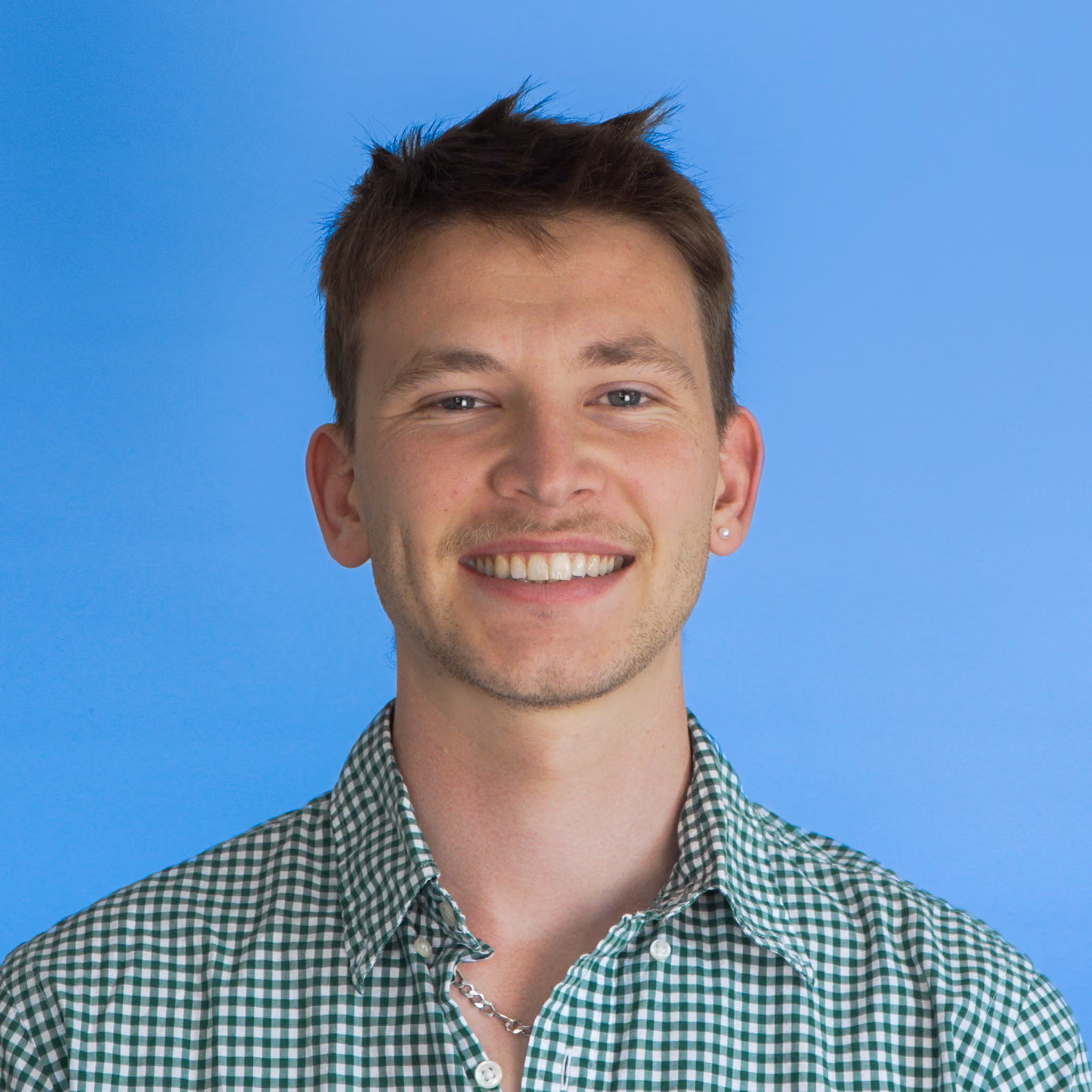 Smiling young man with short brown hair wearing a green and white checkered shirt and a silver chain necklace against a blue background.