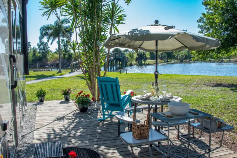 Patio by a lake with chairs, table set for dining, umbrella, and greenery under clear sky.
