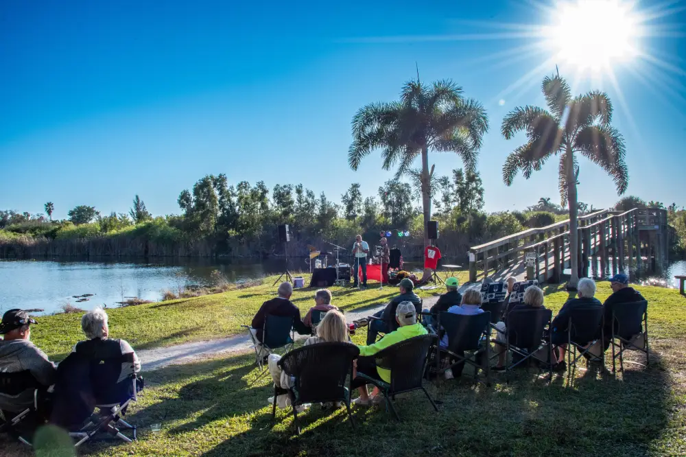 People seated outdoors on chairs by a lake watching a small stage with speakers and performers under bright sun.
