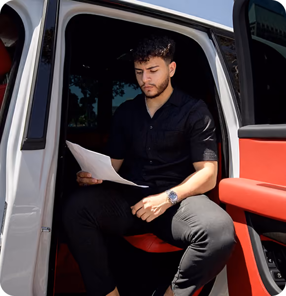 Man in black shirt sitting inside open car door, reading a paper with red car interior.