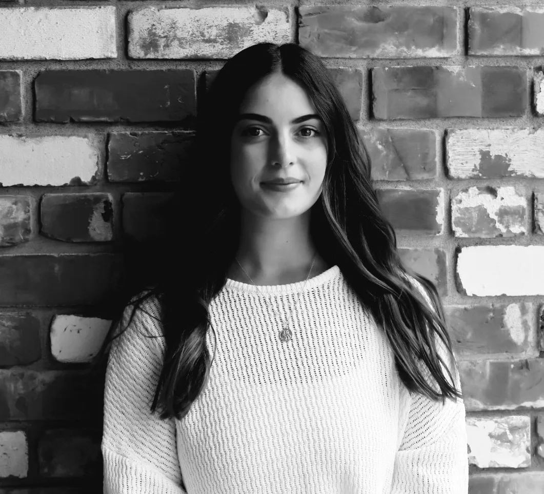 Young woman with long hair in white knit sweater standing against a brick wall, looking at the camera.