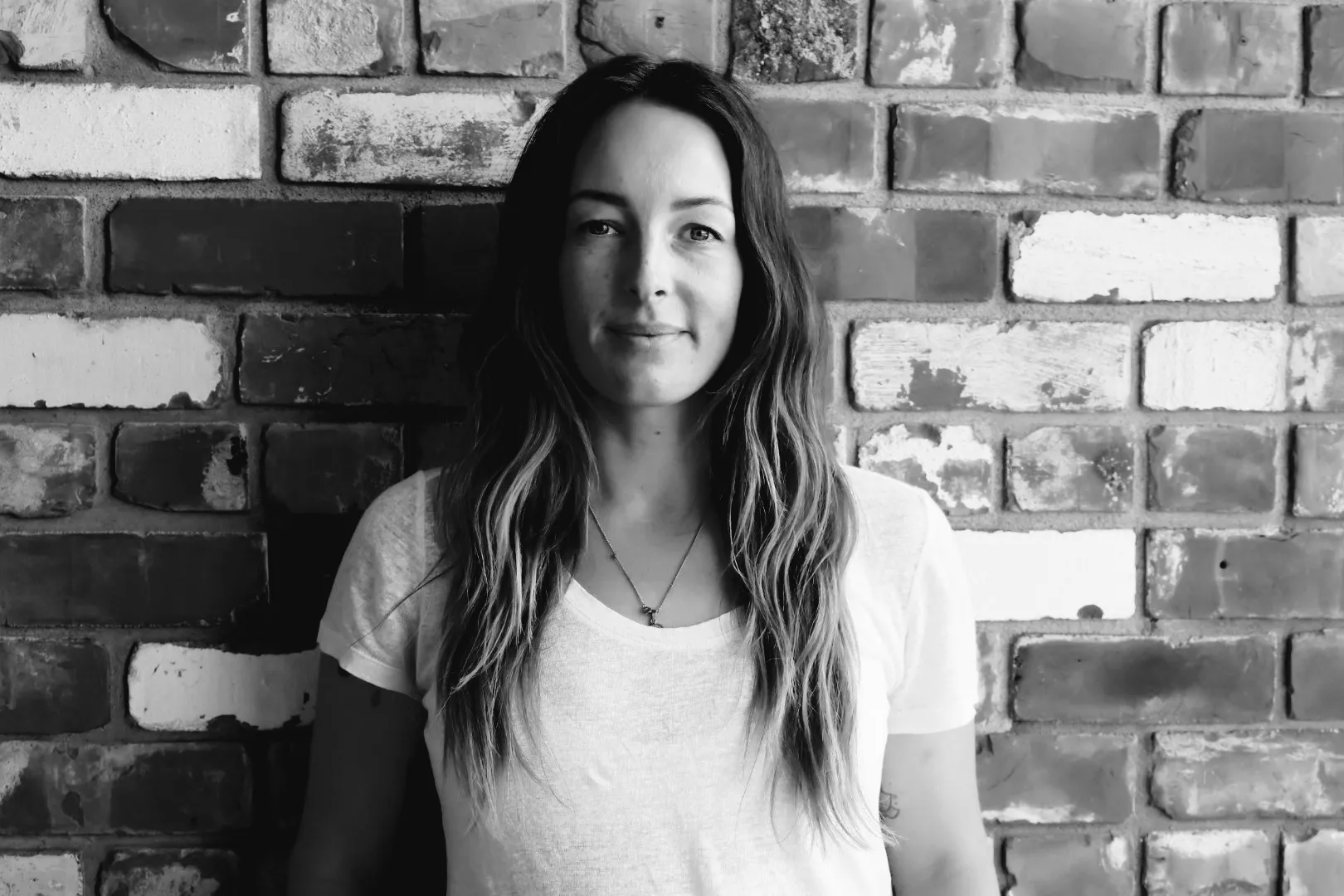 Black and white portrait of a woman with long hair standing against a brick wall background.