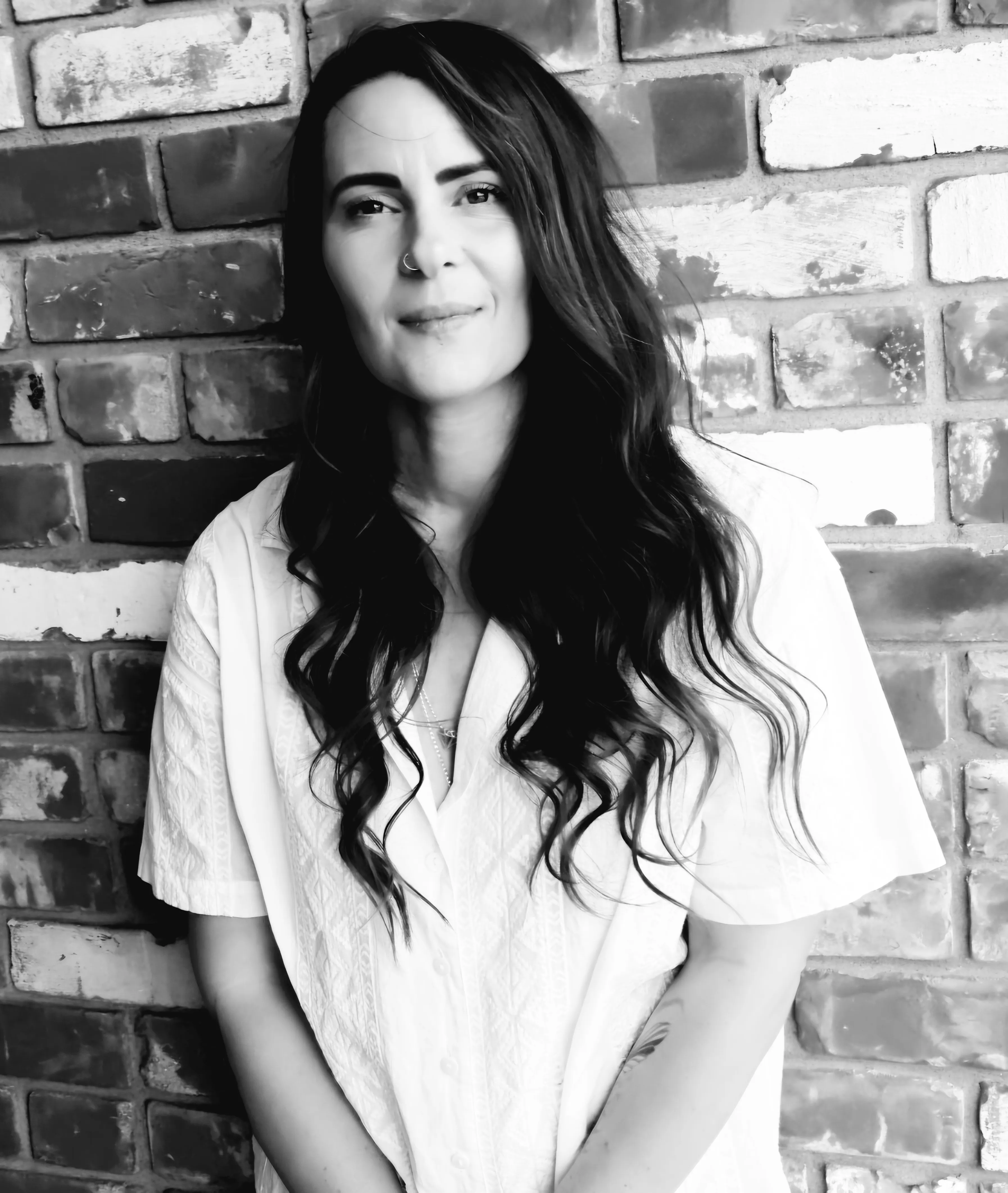 Black and white portrait of a woman with long wavy hair in a white shirt standing against a brick wall.
