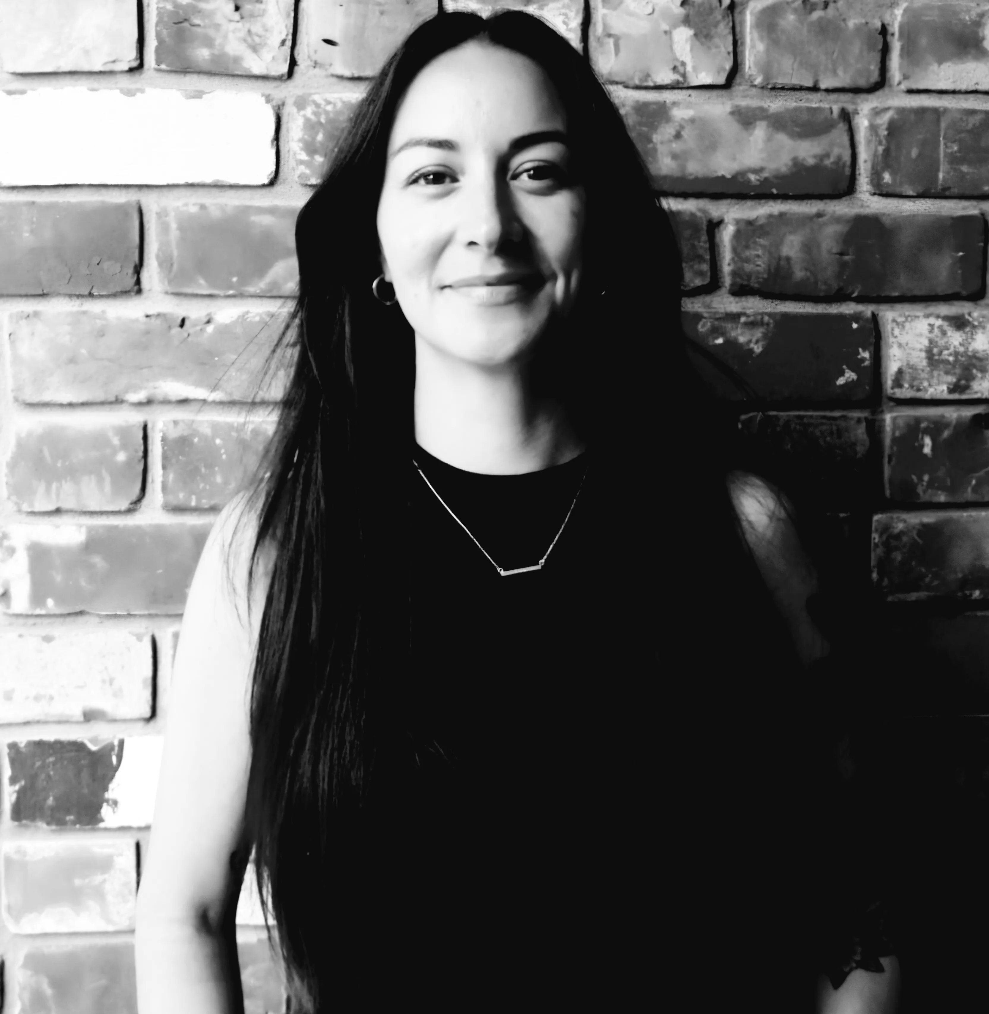 Smiling woman with long dark hair and hoop earrings standing against a brick wall background.