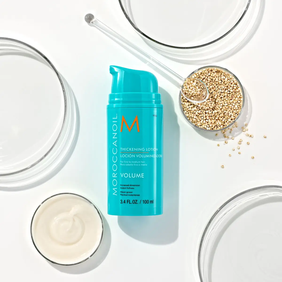 Blue bottle of Moroccanoil Thickening Lotion with bowls of cream, seeds, and glassware on white background.
