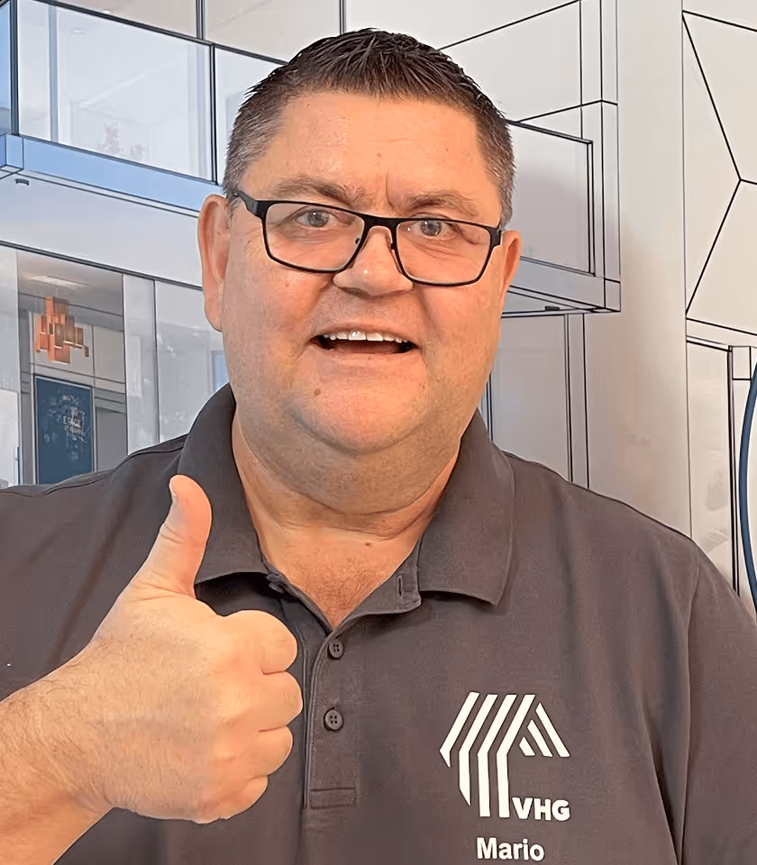 Mann mit Brille zeigt Daumen hoch, trägt graues VHG-Poloshirt, lächelt vor moderner Gebäudezeichnung im Hintergrund.