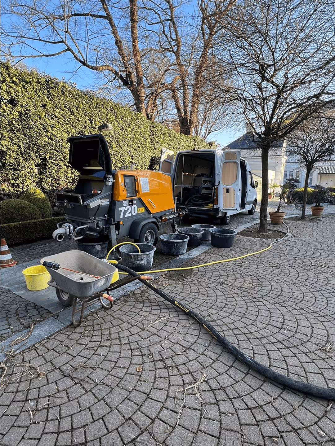 Gelber Betonmischer und weißer Lieferwagen auf gepflastertem Gehweg, umgeben von Eimern, Schubkarre und winterlichen Bäumen.