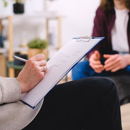 Person writing notes on a clipboard during a conversation with another person sitting nearby.