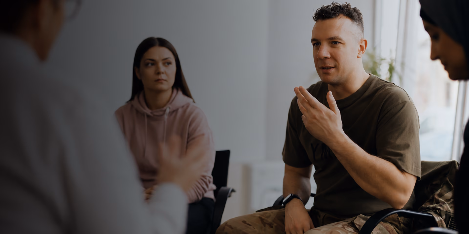 Man in military pants talks while seated with two women in a casual indoor setting.