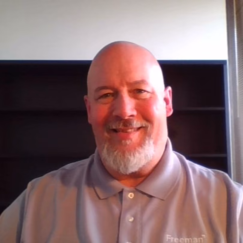 Smiling bald man with a white beard wearing a gray polo shirt sitting before empty dark shelves.