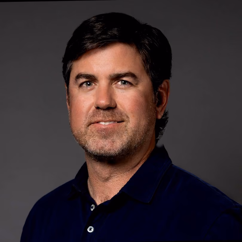 Man with dark hair, slight beard, wearing a dark blue collared shirt against a gray background.