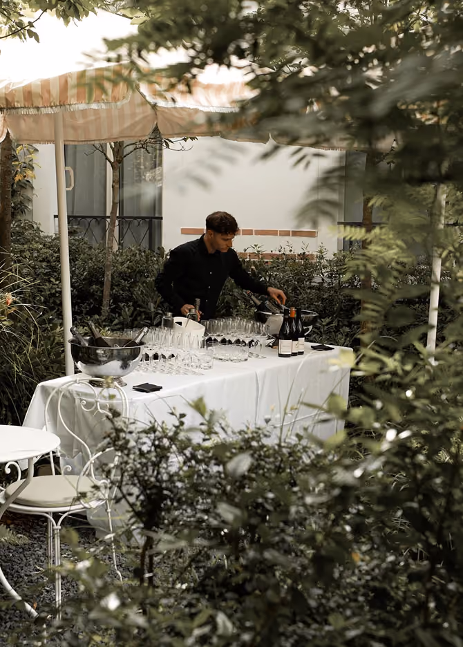Serveur préparant des boissons sur une table dressée dans un jardin verdoyant, sous un parasol rayé