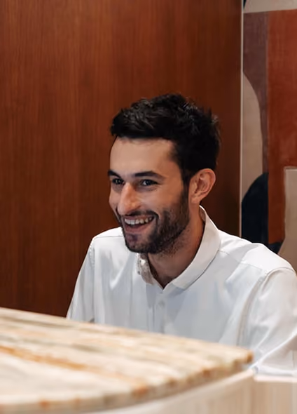 Portrait souriant d'un jeune homme brun en chemise blanche, derrière un comptoir en marbre et boiseries chaleureuses