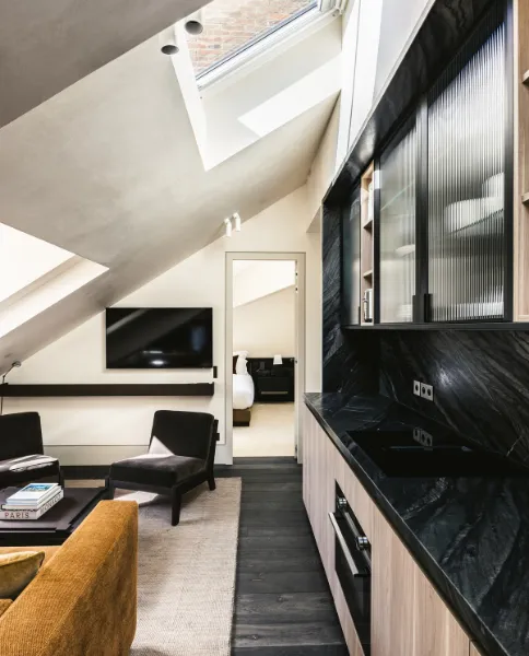 Suite mansardée avec verrière au plafond, kitchenette en marbre noir et salon avec canapé moutarde et fauteuils noirs