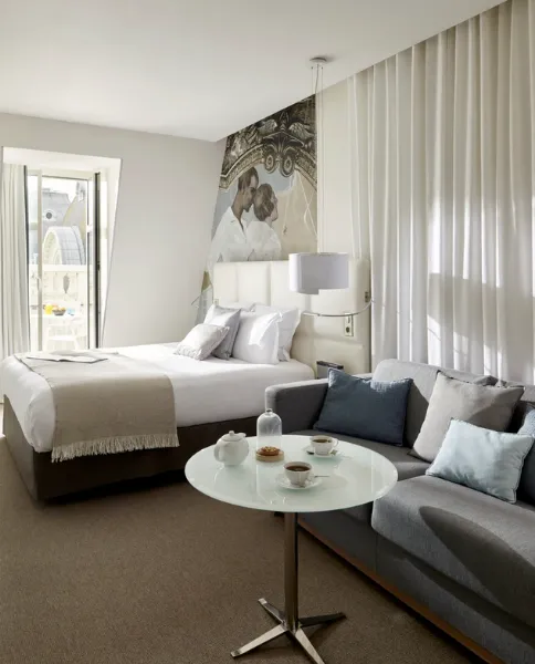 Chambre d'hôtel avec lit blanc, canapé gris, table basse ronde et fresque murale, vue sur balcon et dôme parisien