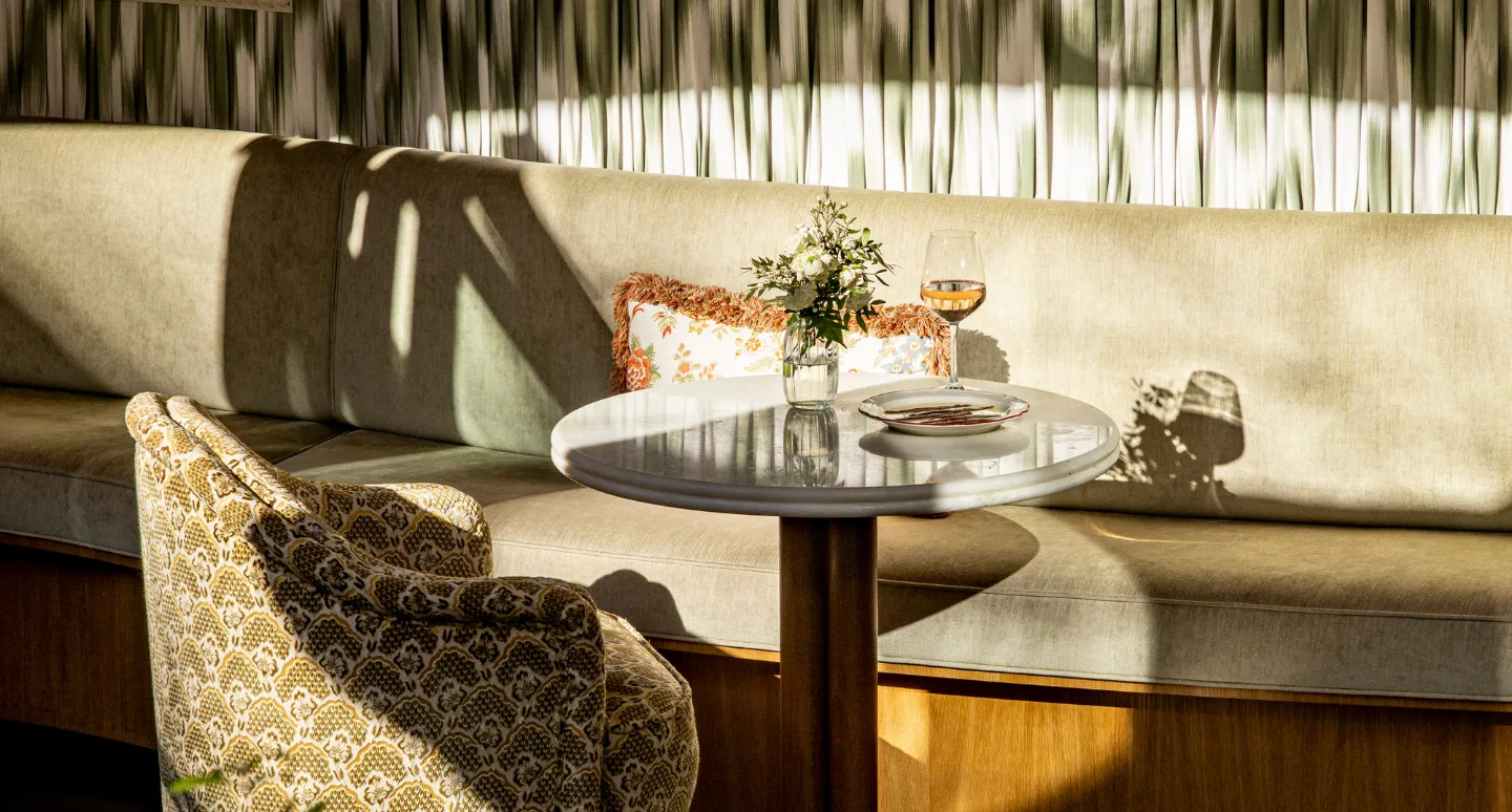 Petite table ronde en marbre avec verre de vin et bouquet de fleurs, banquette beige baignée de lumière tamisée
