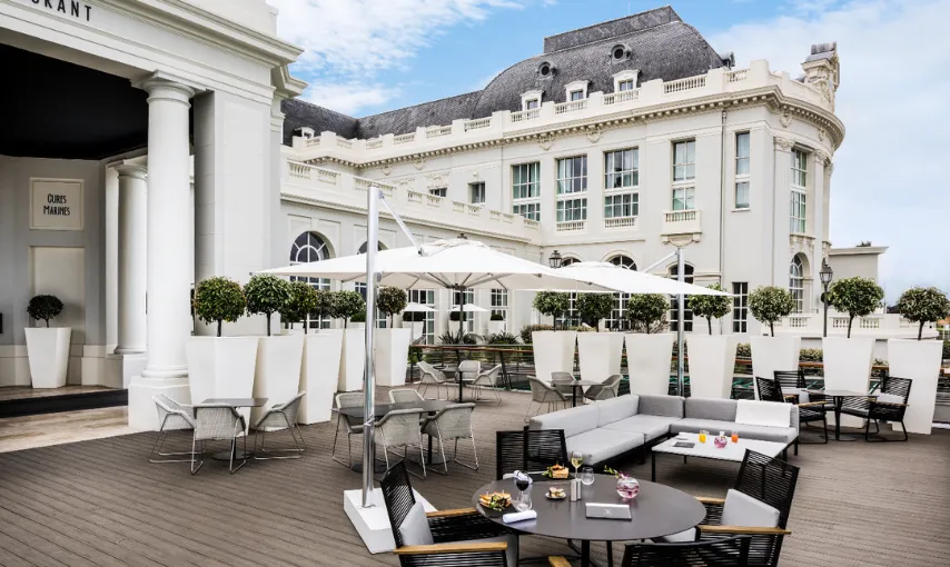 A restaurant terrace with white parasols in front of an elegant hotel façade