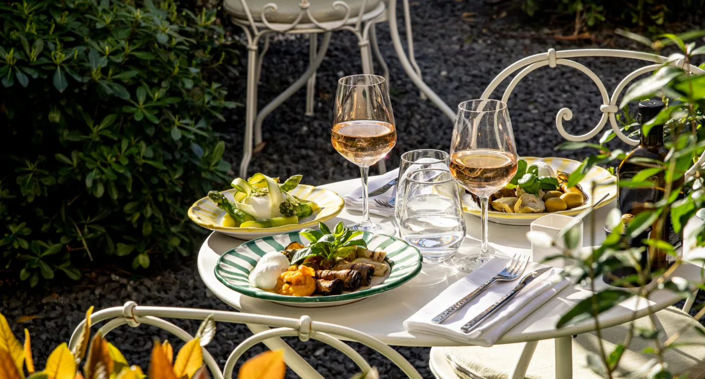 Table de jardin avec assiettes de légumes grillés et burrata, accompagnées de verres de vin rosé au soleil