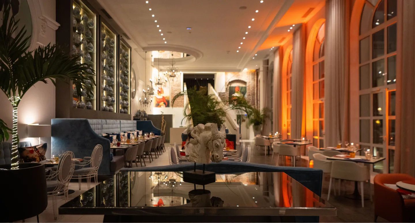 Salle de restaurant nocturne avec banquette bleue, sculpture de cheval et grandes arcades éclairées d'orange