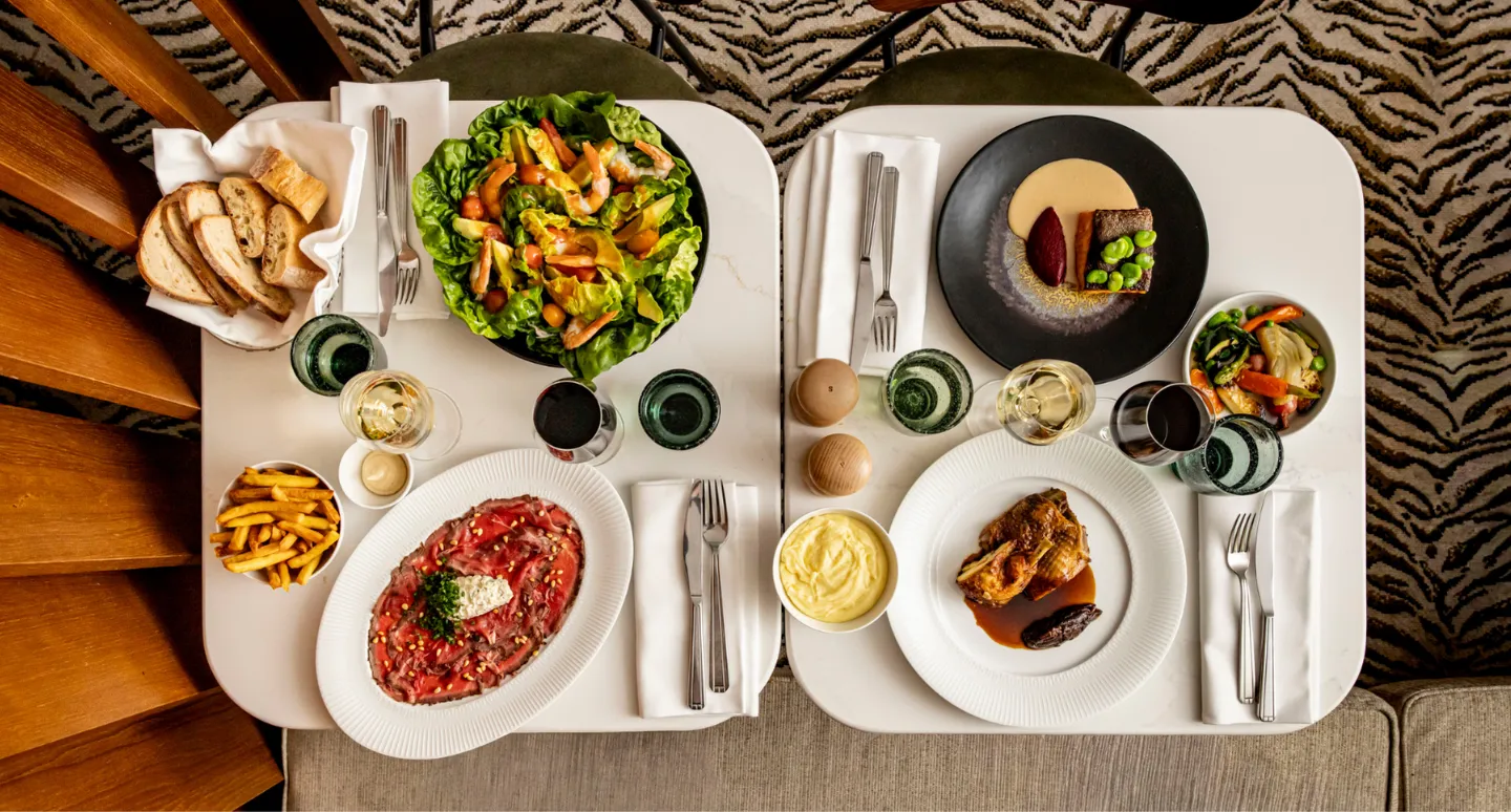 Vue aérienne de deux tables dressées avec carpaccio, salade, poulet rôti et dessert, accompagnés de verres de vin