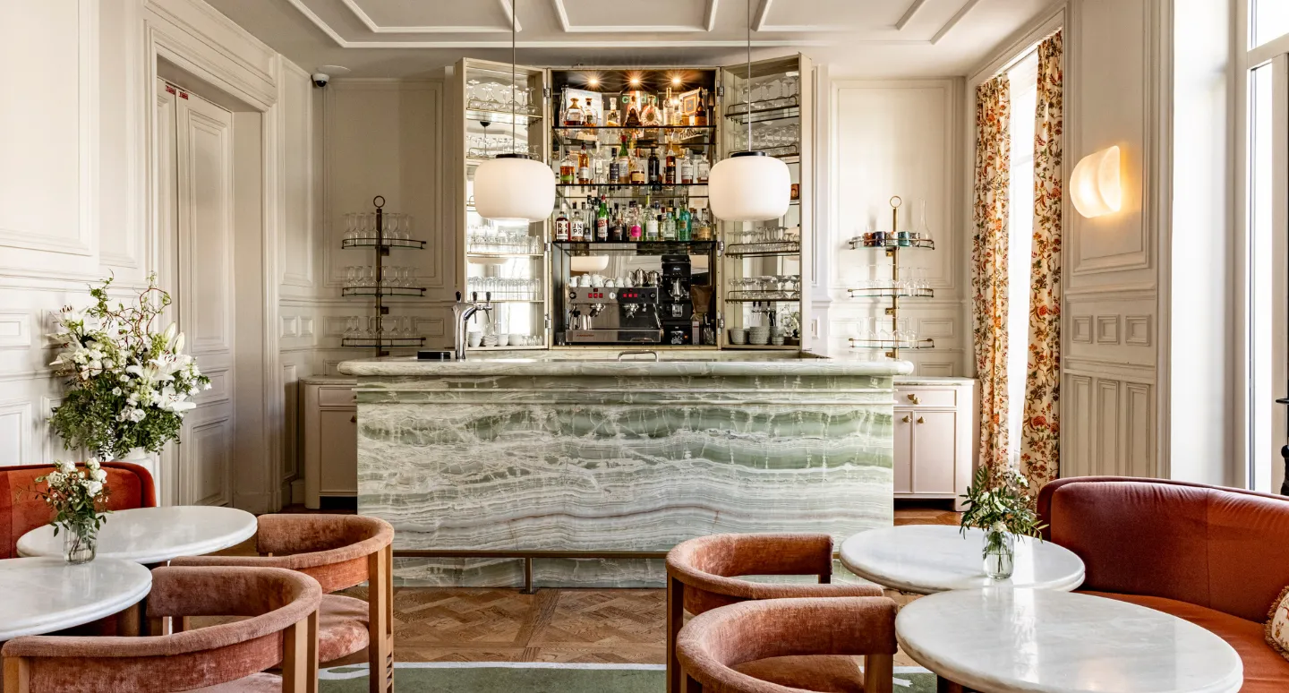 Bar en marbre vert veiné avec étagères à bouteilles, entouré de fauteuils et tables rondes en velours orange