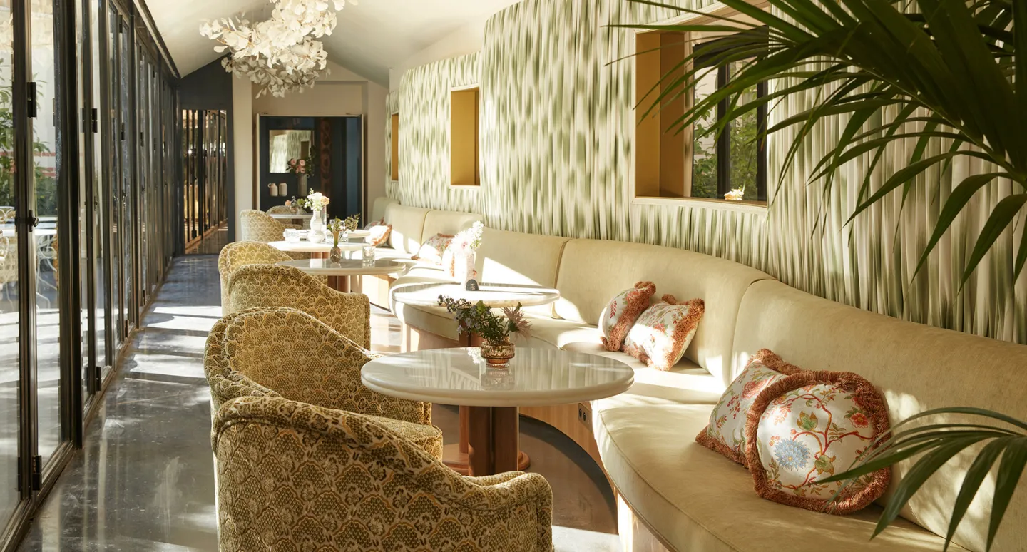 Salon lumineux avec banquette beige incurvée, fauteuils à motifs et lustre floral, verrière ouverte sur un couloir vitré