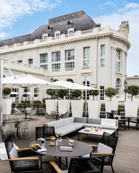 An outdoor terrace with sofas and tables overlooking the majestic façade of the Cures Marines hotel