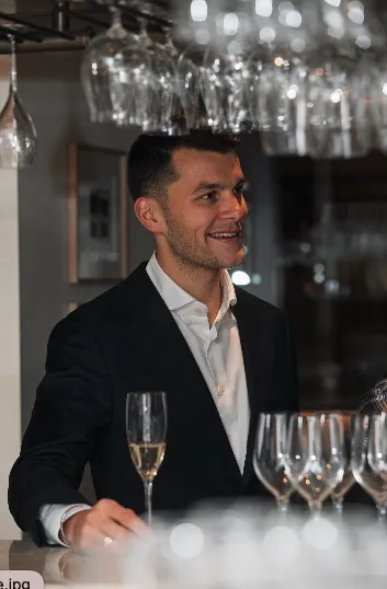A smiling man in a dark suit behind a bar, surrounded by hanging wine glasses and a champagne flute