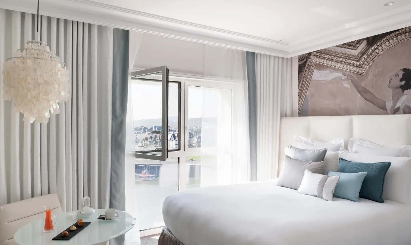Chambre d'hôtel avec lit blanc, lustre en coquillages et fenêtre ouverte sur le port de Trouville-sur-Mer