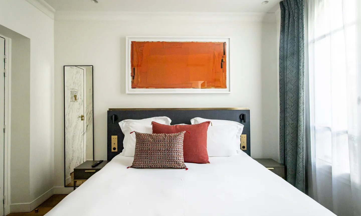 Chambre d'hôtel épurée avec lit blanc, tête de lit noire, coussins rouges et tableau abstrait orange au mur