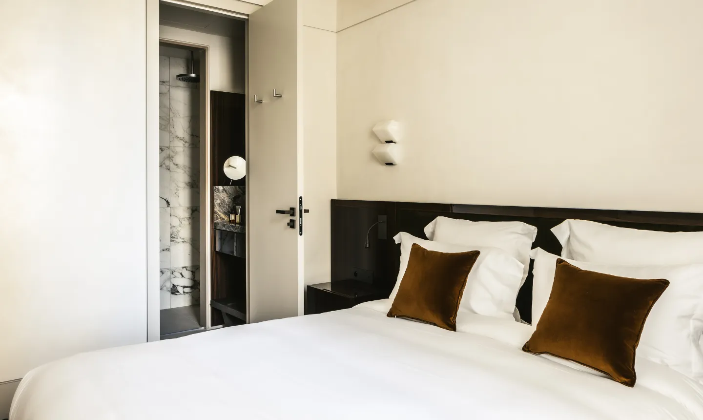 Chambre d'hôtel avec lit blanc et coussins en velours marron, vue sur salle de bain en marbre
