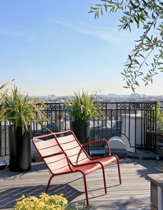 Terrasse rooftop avec chaise longue rouge et jardinières verdoyantes, vue sur les toits de Paris