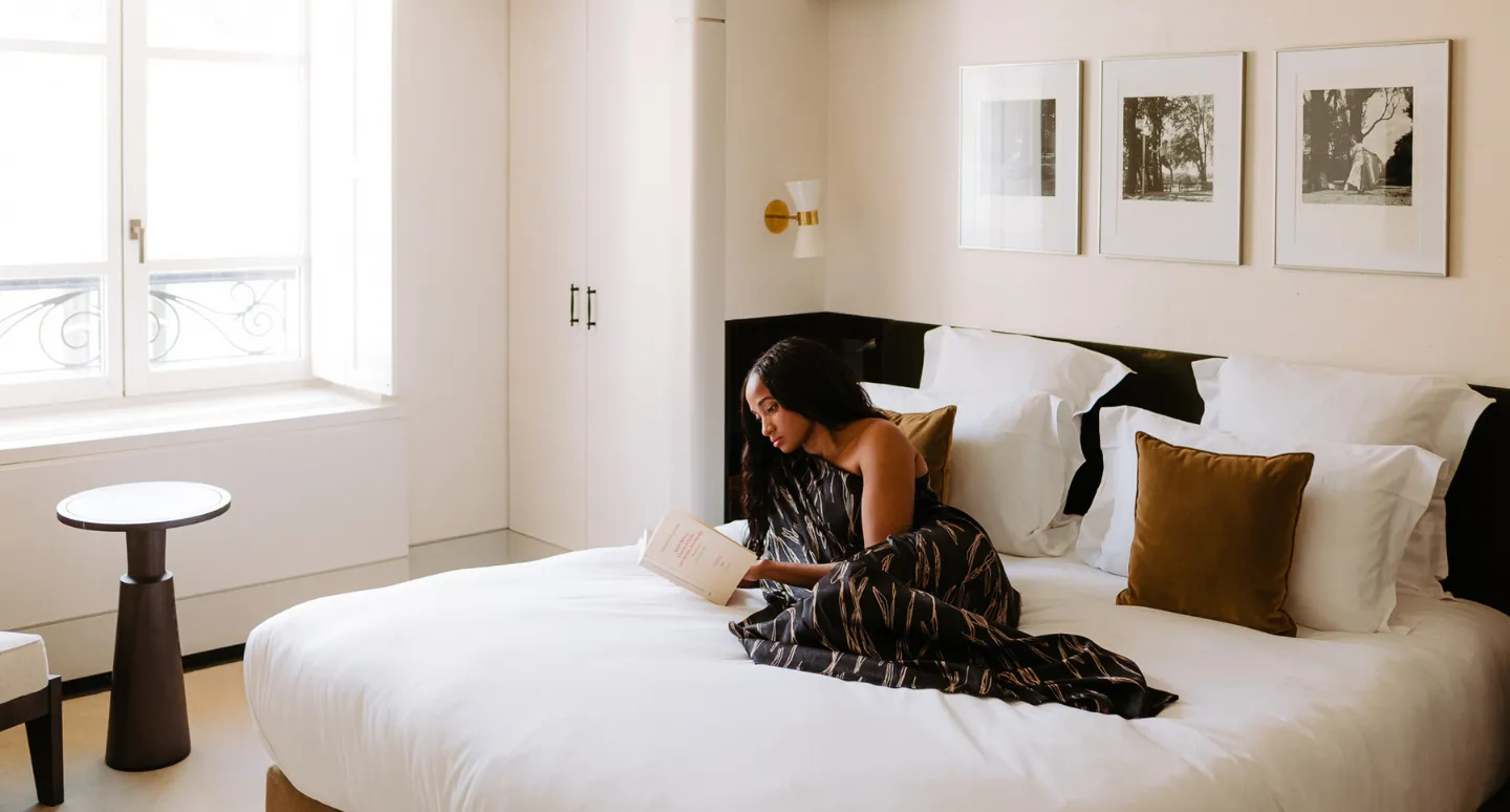 Femme lisant un livre sur un lit blanc, enveloppée d'un tissu imprimé, dans une chambre lumineuse aux photographies encadrées
