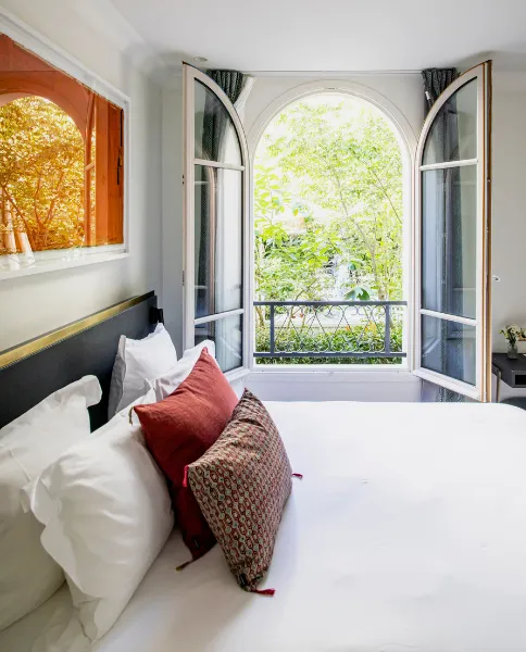 Chambre lumineuse avec lit à coussins rouges et fenêtre arquée ouverte sur un balcon fleuri et arboré