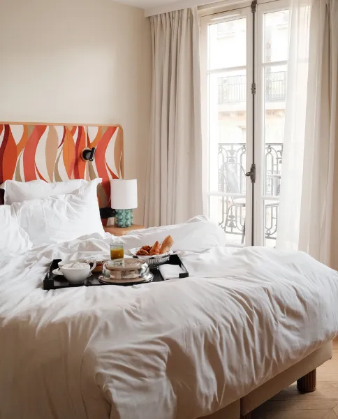 Chambre lumineuse avec lit blanc, tableau floral coloré au mur et fenêtres donnant sur des balcons parisiens