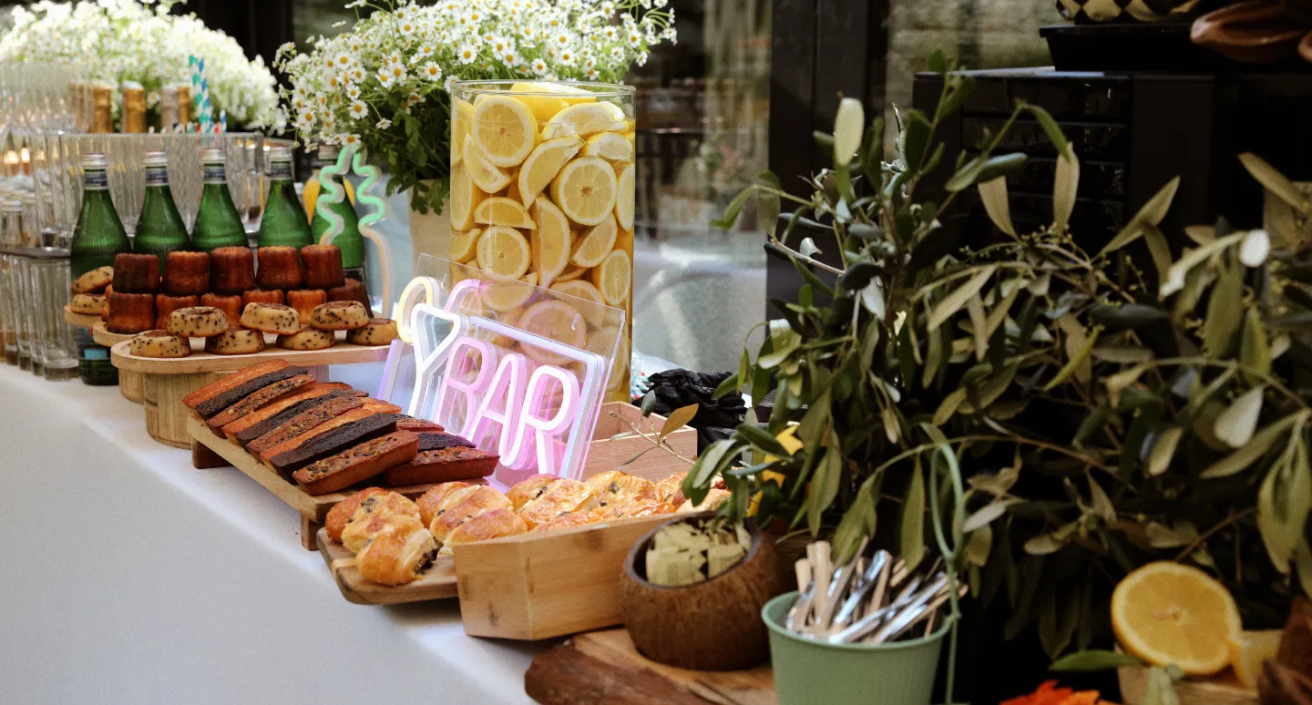 Buffet de brunch avec viennoiseries, cannelés et citrons, enseigne néon Bar rose et décor floral d'olivier
