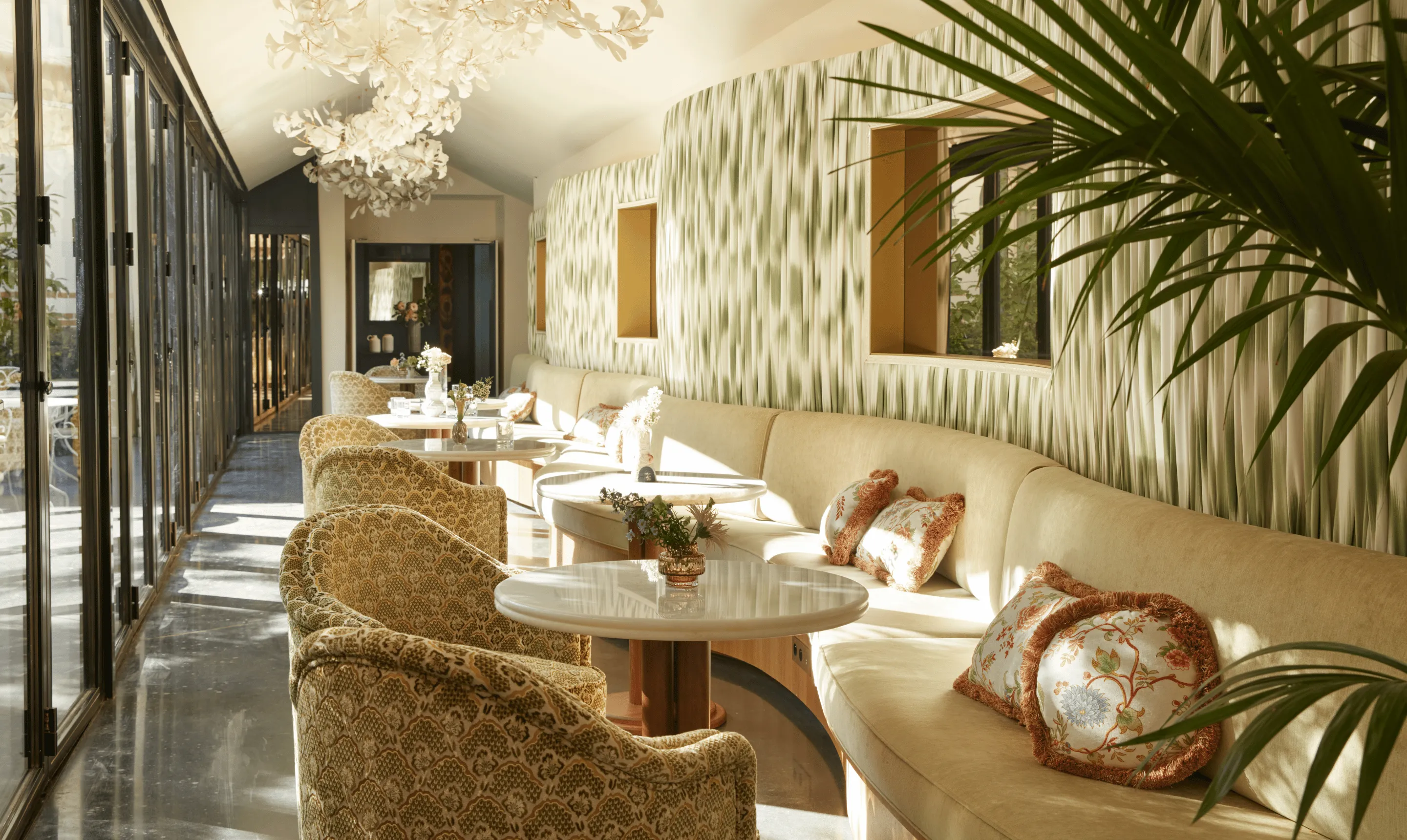 Salon lumineux avec banquette beige incurvée, fauteuils à motifs, tables de marbre et lustre floral, baie vitrée sur jardin