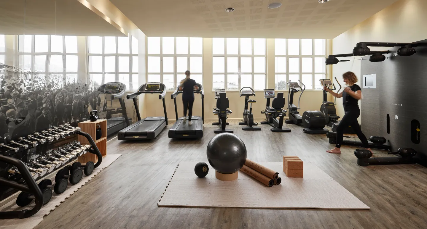 Salle de fitness lumineuse avec tapis de course, vélos, haltères et deux personnes s'entraînant devant de grandes fenêtres