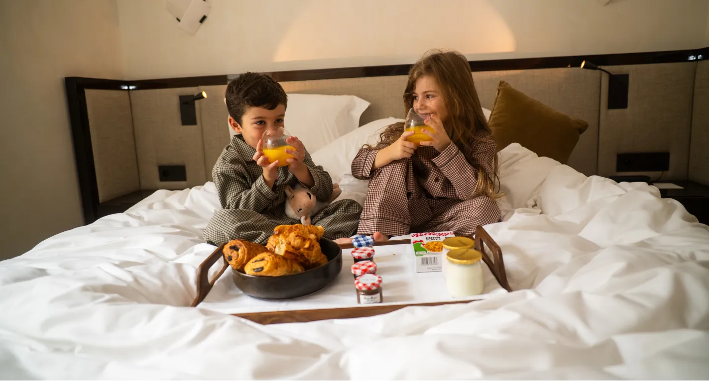 Deux enfants en pyjama buvant du jus d'orange sur un lit d'hôtel, devant un plateau de viennoiseries et confitures