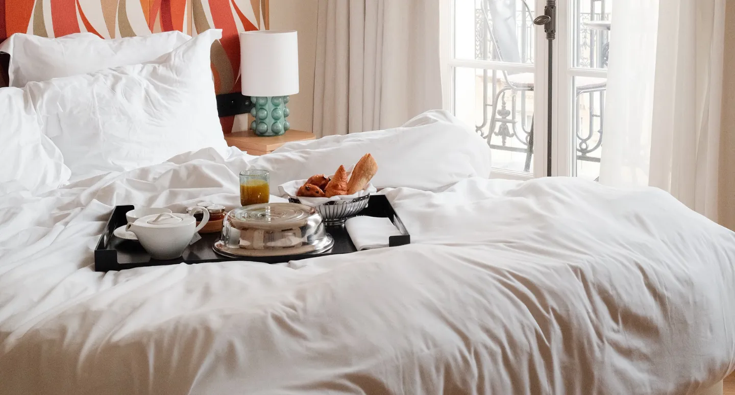 Plateau petit-déjeuner avec théière, viennoiseries et jus de fruit servi sur un lit blanc face à un balcon parisien