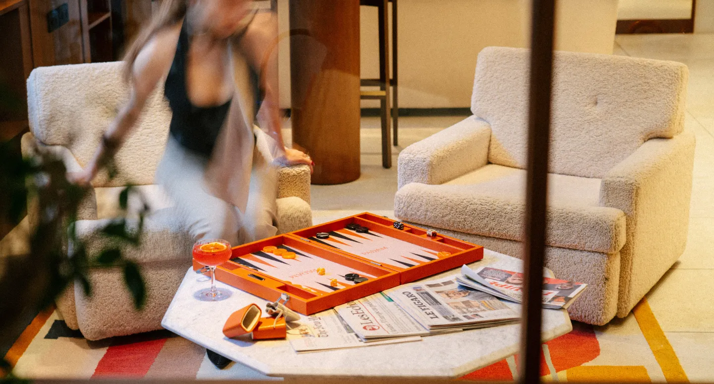 Jeu de backgammon orange posé sur une table basse en marbre, entre fauteuils en bouclette blanche, avec cocktail et journaux